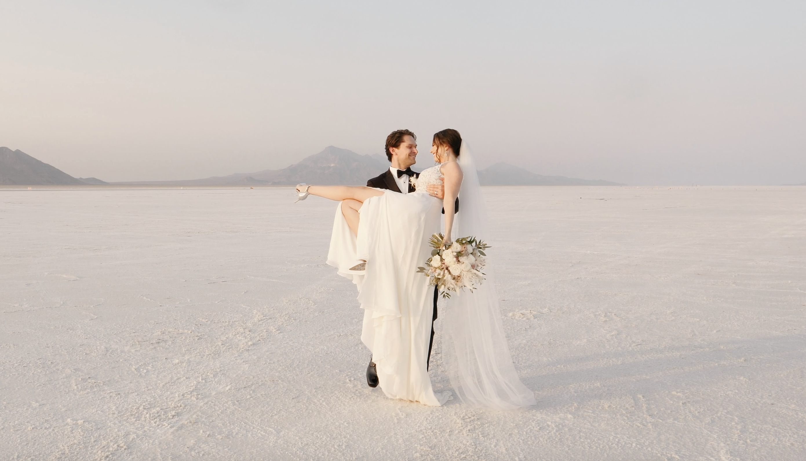 bride and groom on utah salt flats, groom is holding bride, bride is in a long, sleeveless white gown. She is wearing a long veil. The groom suit is the classic black.